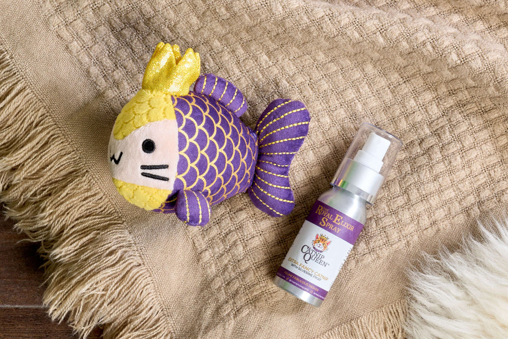 Purple and gold plush fish toy with yellow crown next to a silver Royal Elixir Spray bottle on a tan woven blanket